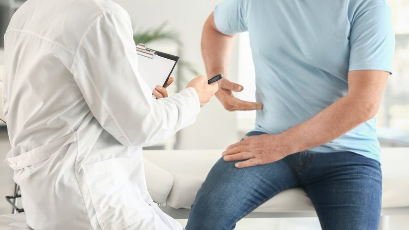 Arzt untersucht einen Patienten mit Rückenschmerzen, der auf der Liege sitzt und auf seine Seite zeigt.