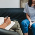 Psychologisches Gespräch: Eine Frau sitzt auf einem Sofa, die Therapeutin macht sich Notizen