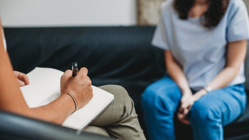Psychologisches Gespräch: Eine Frau sitzt auf einem Sofa, die Therapeutin macht sich Notizen